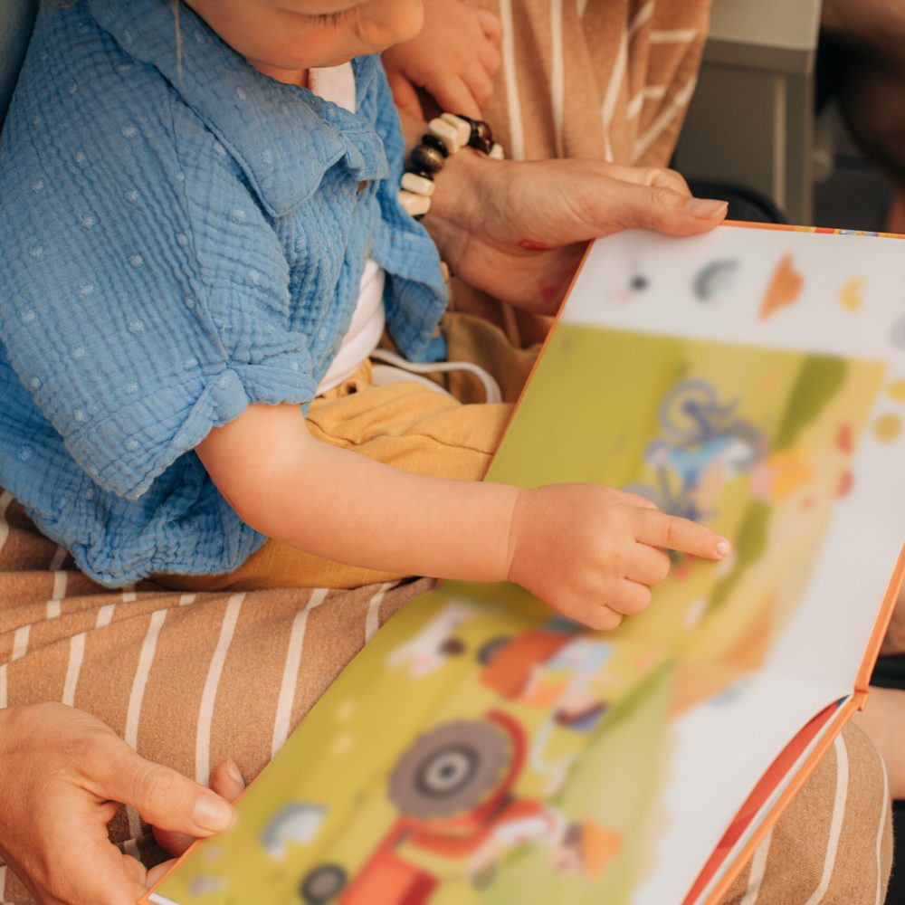 Cute little girl pointing at pictures in book. Toddler sitting on her mother laps and reading book. Family at home, child development concept